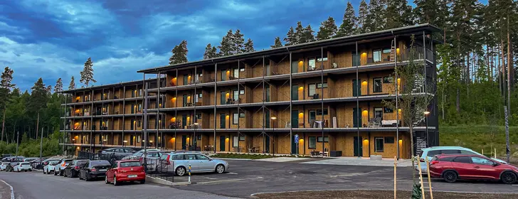 Flerbostadshus med brun träfasad, svarta fönster samt dörrar, parkering på framsidan och skog på baksidan | Nivika Fastigheter AB (publ)