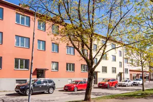 Flerbostadshus med gul, rosa och vit putsad fasad, träd i förgrunden | Nivika Fastigheter AB (publ)