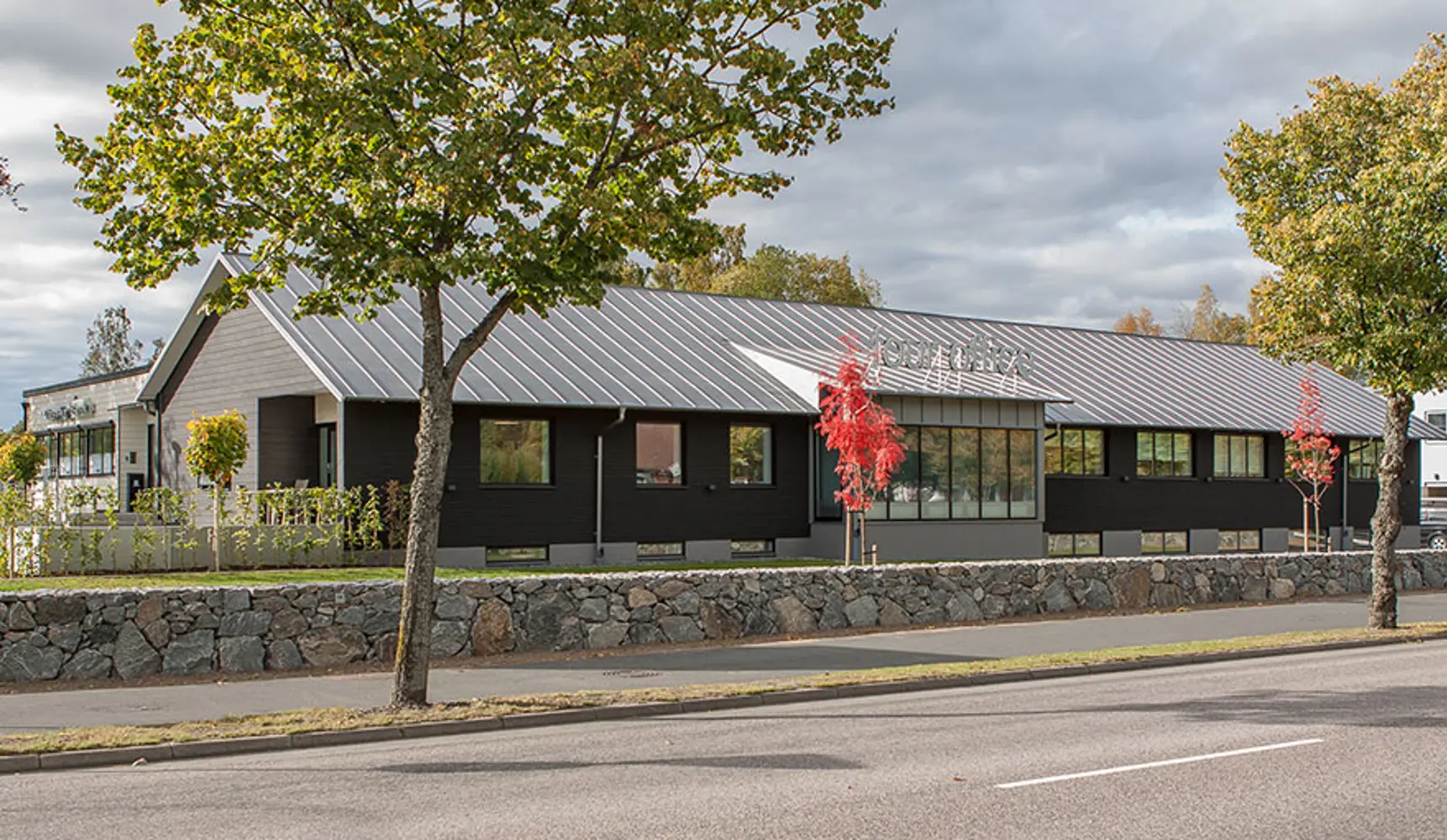 Kommersiell fastighet med svart fasad, svarta fönster samt svart tak, stenmur i förgrunden | Nivika Fastigheter AB (publ)